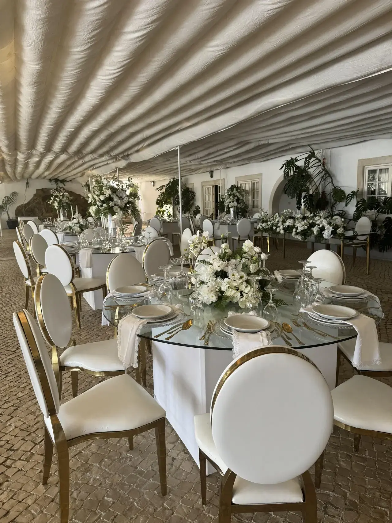 Salão interior da quinta com longa mesa de banquete, cadeiras brancas e douradas